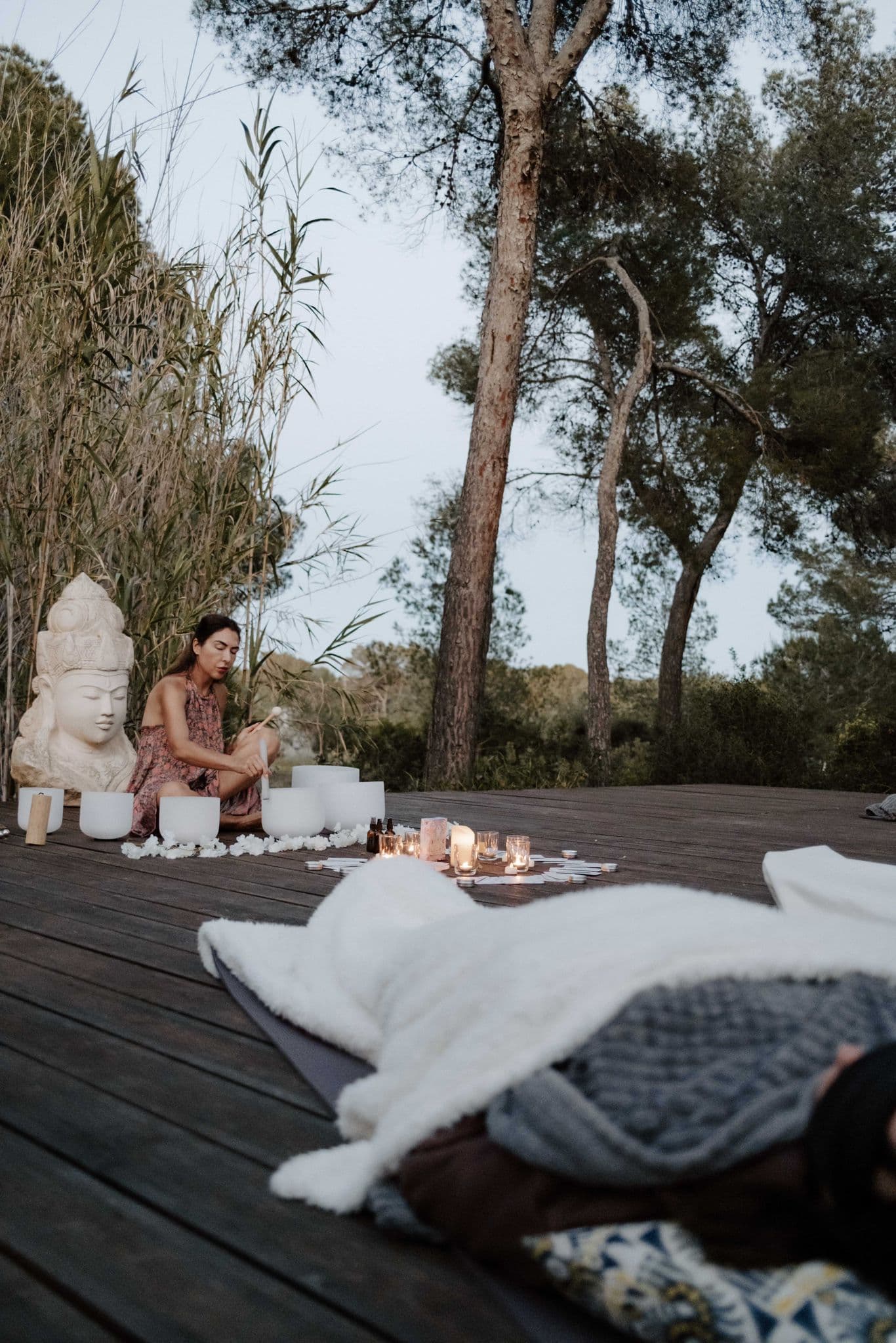 Tanya seated by a stone deity at twilight, surrounded by crystal singing bowls and candles in Ibiza